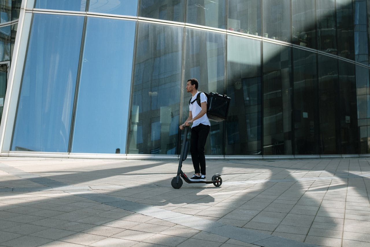 Man on electric scooter with backpack in front of modern glass building, representing urban delivery service.